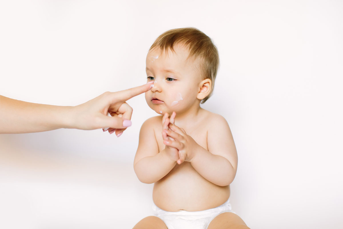 Baby Gesicht wird von Mutter eingecremt und Baby klatscht vor Freude in die Hände als Sinnbild für die richtige Hautpflege bei Neugeborenen