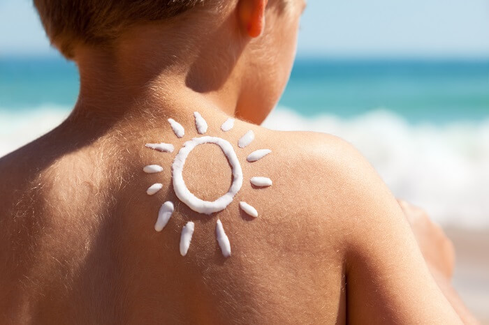 Kind sitzt am Strand und hat eine Sonne aus Sonnencreme auf den Rücken gemalt als Sinnbild für Sonnenschutz für Babys und Kinder