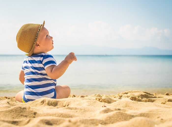 Rückansicht eines Babys mit Sonnenhut, das am im Ringelbody am Sandstrand sitzt